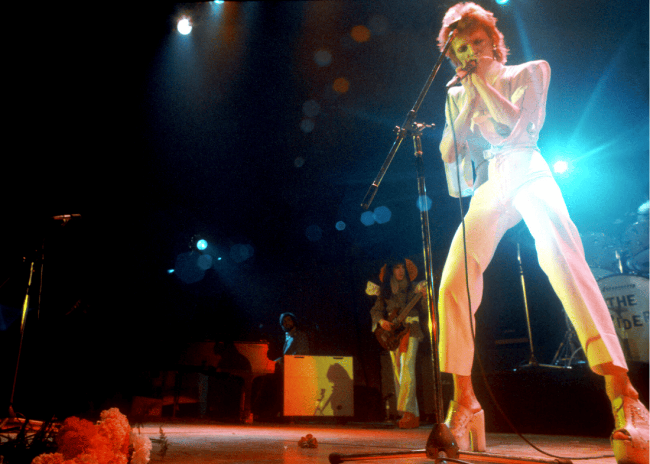 Bowie in a tight white suit with platform heels performs on stage.