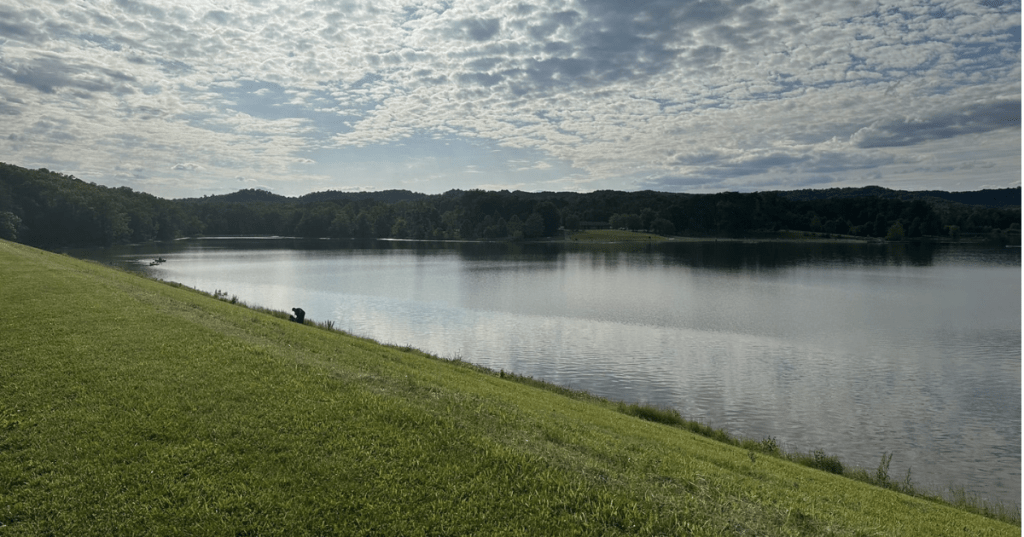 Scenic view of Deam Lake, Indiana from a grassy hill under a bright, cloudy afternoon sky.