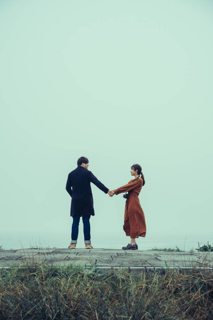 Two people stand on a foggy platform, holding hands and facing each other. One wears a long coat; the other wears a flowing dress