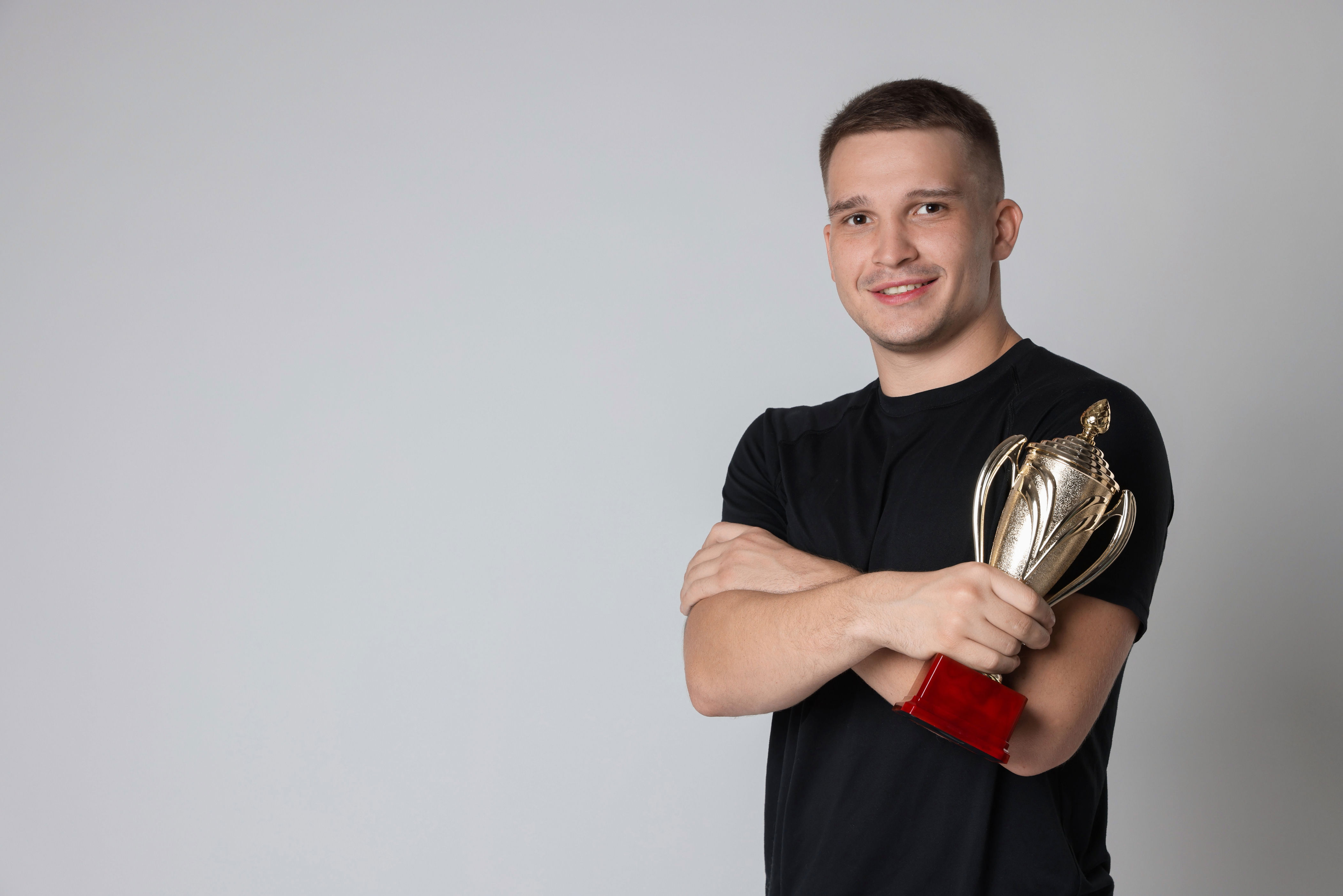 Person in a black t-shirt holding a trophy and smiling against a plain background