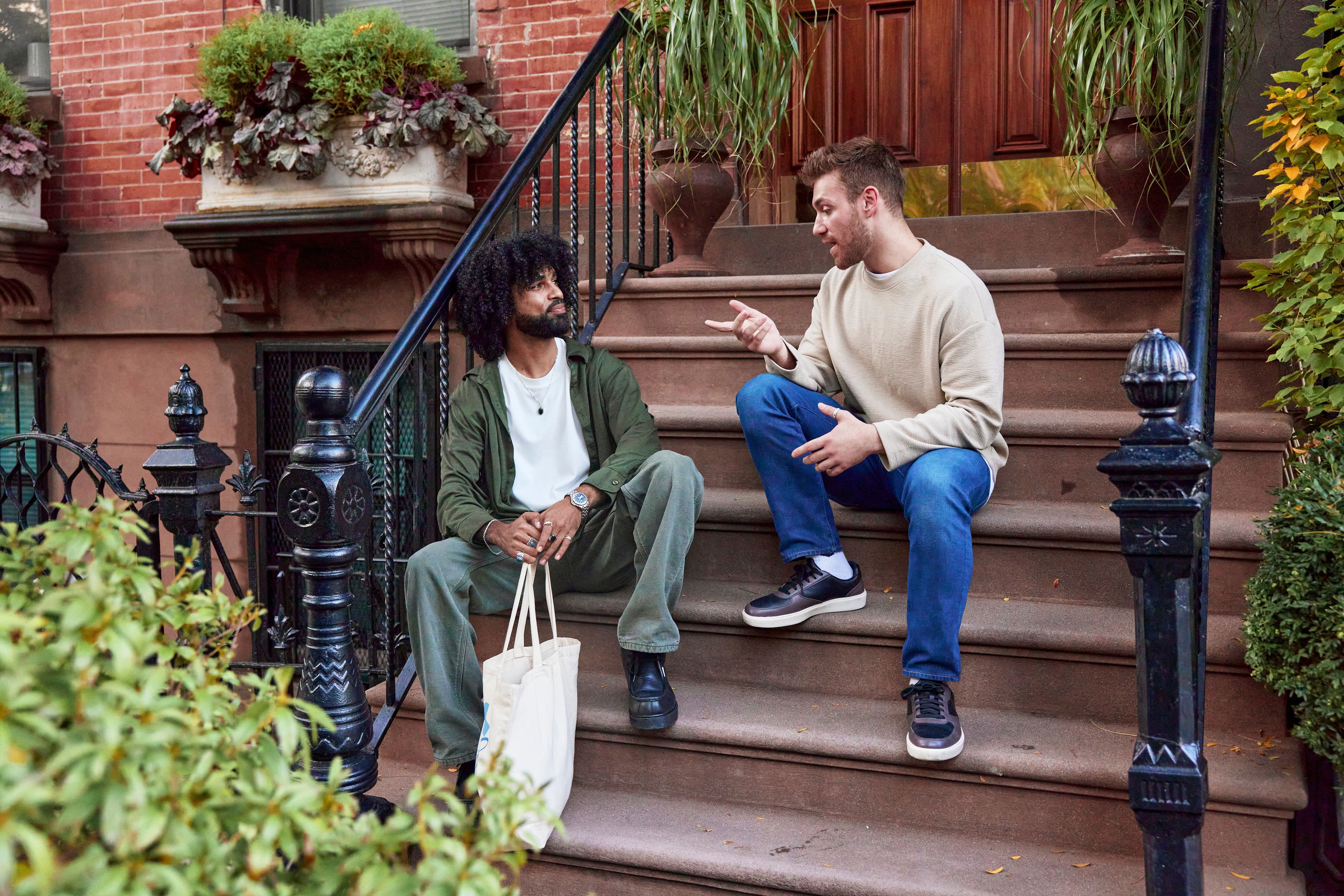 Two people sit on outdoor steps having a casual conversation. One wears a casual green outfit with a tote bag, the other wears jeans and a beige sweater