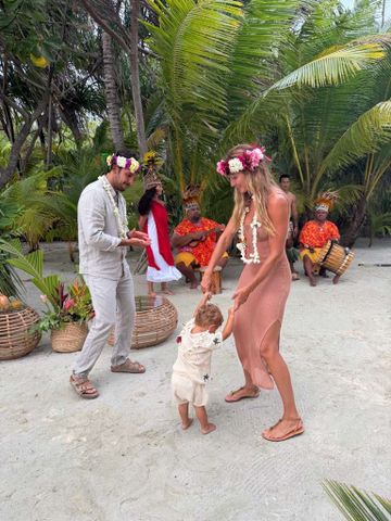 Gisele Bündchen and Joaquim Valente dance on the beach with their one-year-old son in front of a grove of palm trees. Valente and Bündchen are both wearing pink-and-white flower crowns and matching white flower necklaces.