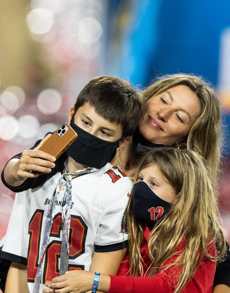 Gisele Bündchen snaps a selfie with her kids, Benjamin and Vivian. The children are wearing coordinated football jerseys and black face masks as Bündchen crouches behind them with a smile.