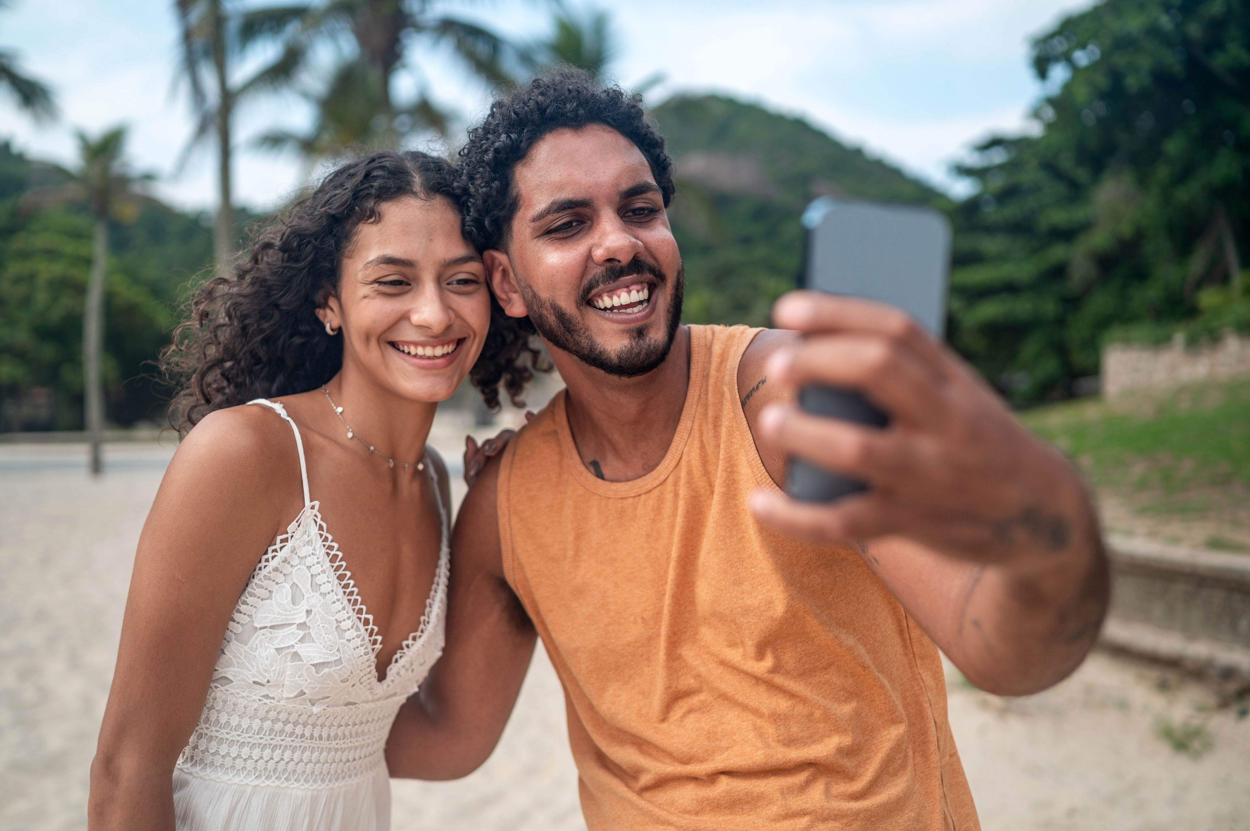 A smiling couple takes a selfie on a beach, with palm trees visible in the background