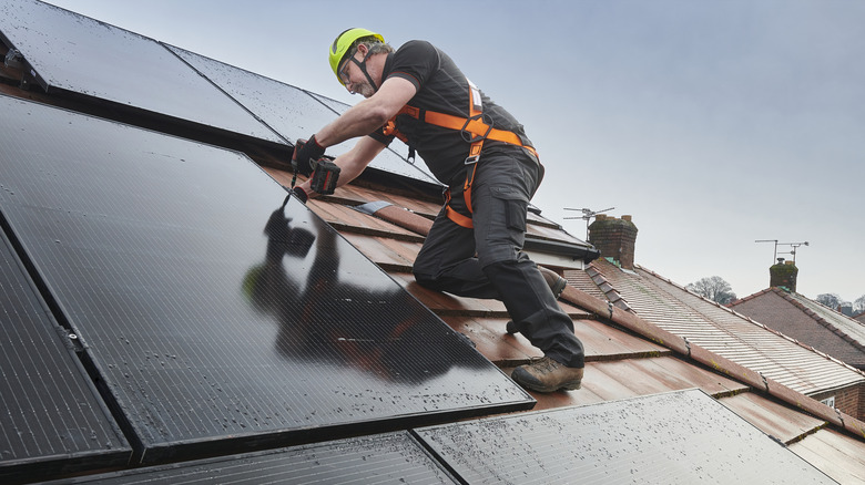 Solar installer securing rooftop panels with a drill while wearing safety gear.
