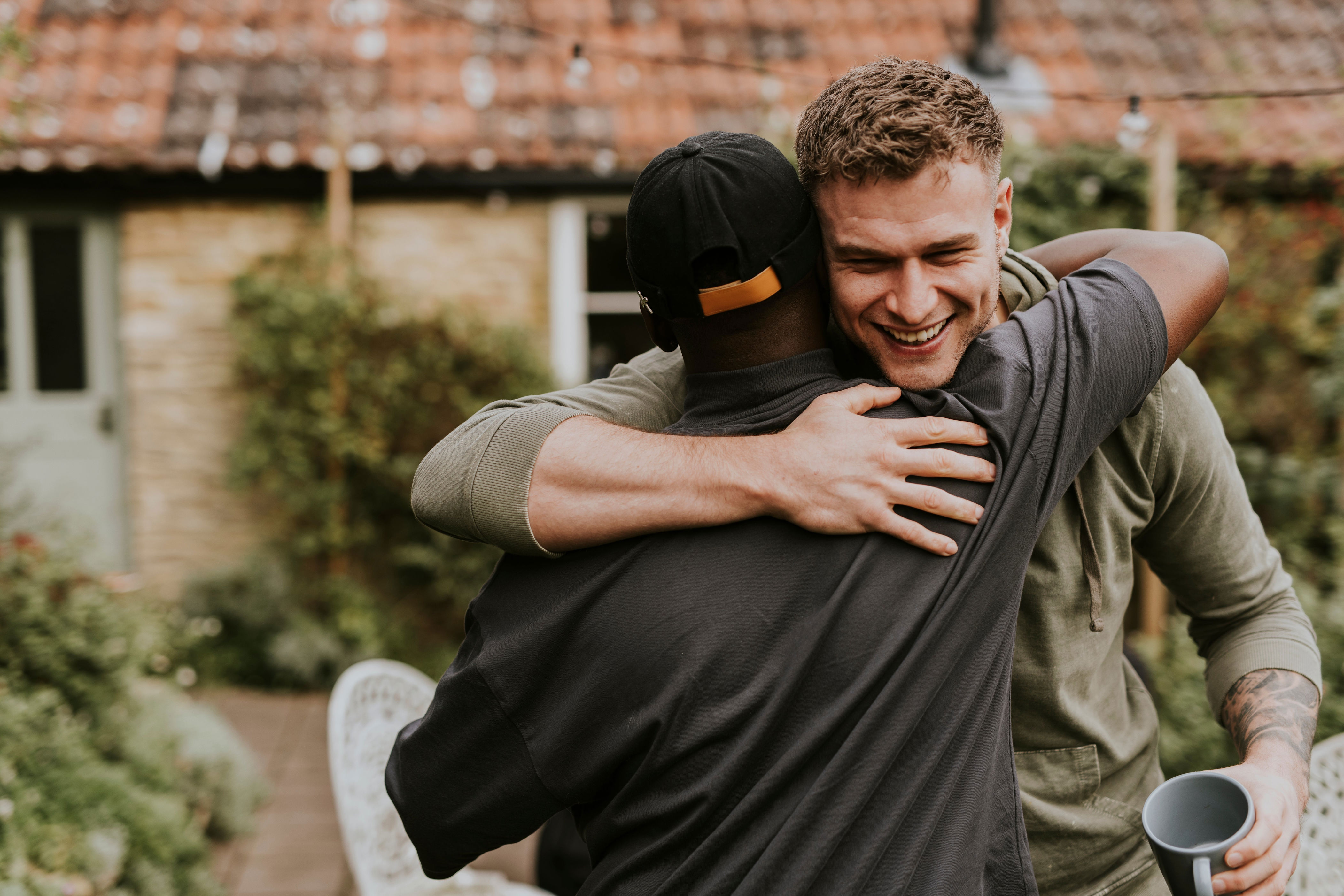 Two friends share a joyful embrace outdoors, one holding a cup. The scene is casual and warm, suggesting a reunion or celebration