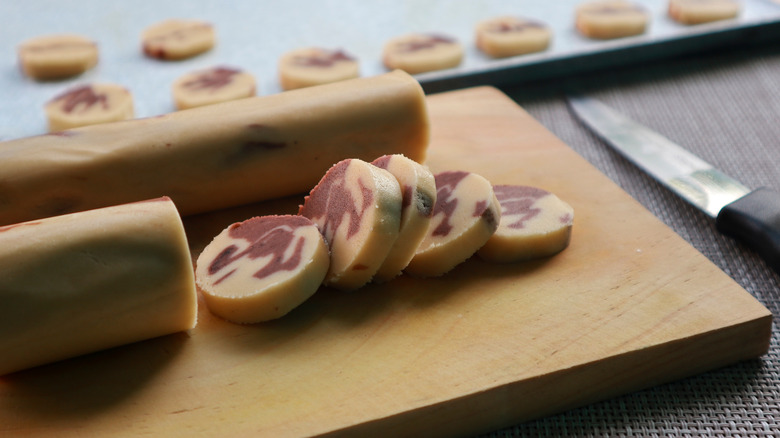 A tube of cookie dough on a cutting board being sliced into uniform medallions and laid out on a baking sheet in the background