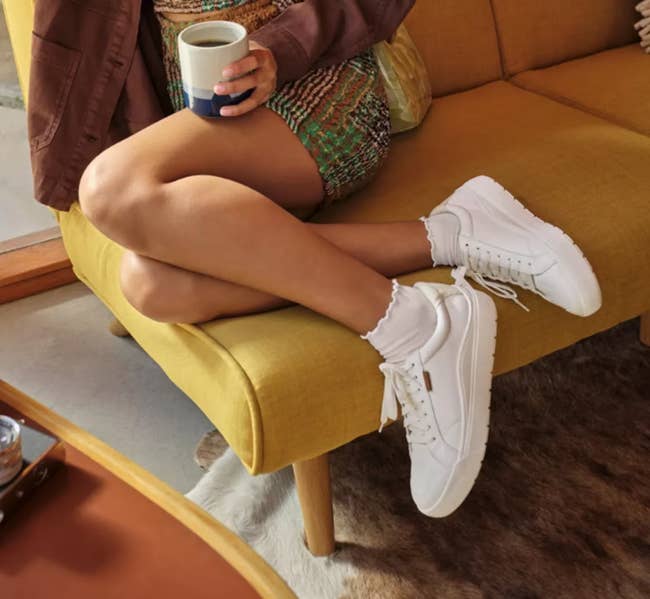 Person in casual outfit, sitting on a couch, holding a mug. White sneakers with prominent branding are the focus. A camera is on the table nearby