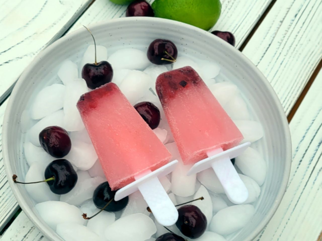 two pink popsicles in a white bowl of ice with cherries on a white painted wooden table