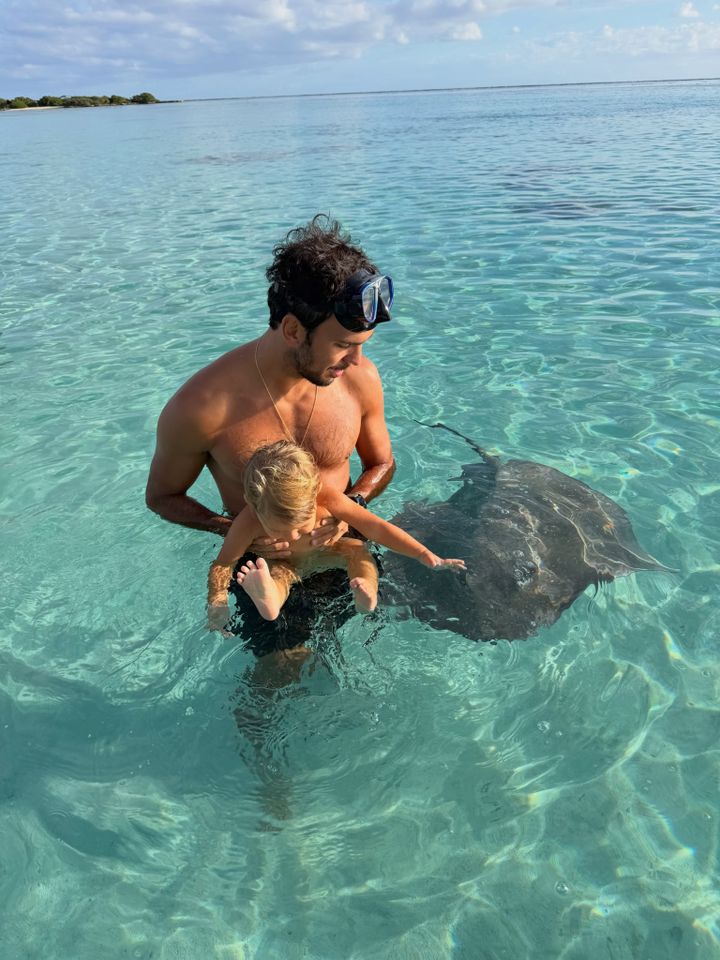Joaquim Valente, Gisele Bündchen's husband, holds their one-year-old baby in the water as they look down at a sting ray in the water. Valente is shirtless and wearing a pair of swim goggles on his forehead.