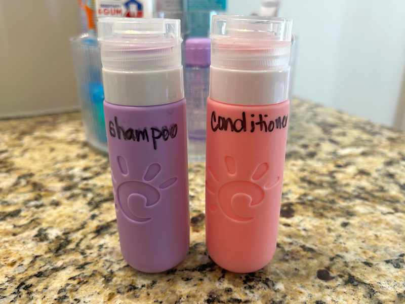 Two labeled travel-sized bottles of shampoo (purple) and conditioner (pink) on a granite countertop.