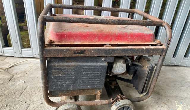 A rusty generator with a red top and black base, sitting on a concrete floor near metal storage.