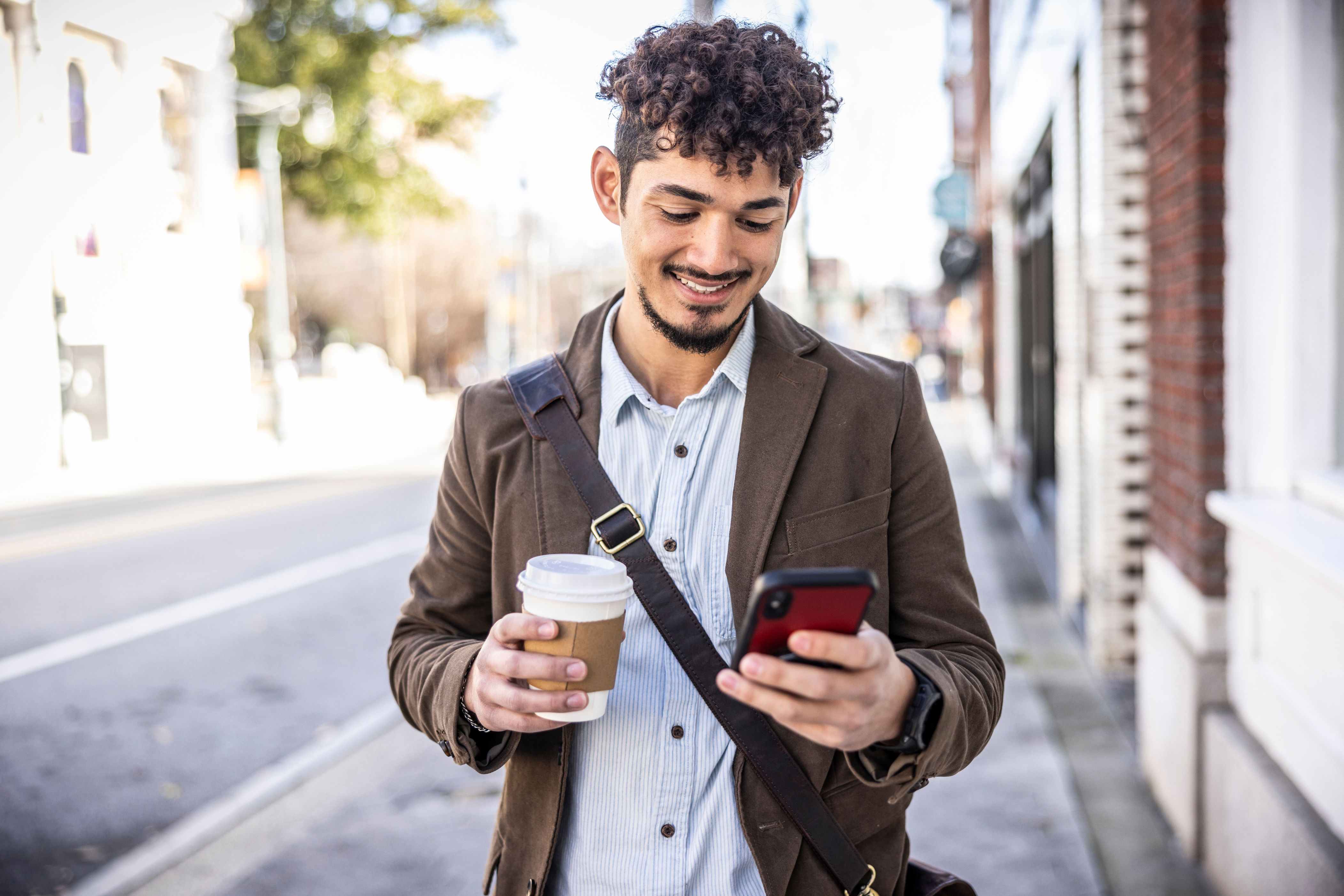 Person smiling while checking their phone, holding a coffee cup, and walking on a city street in casual attire