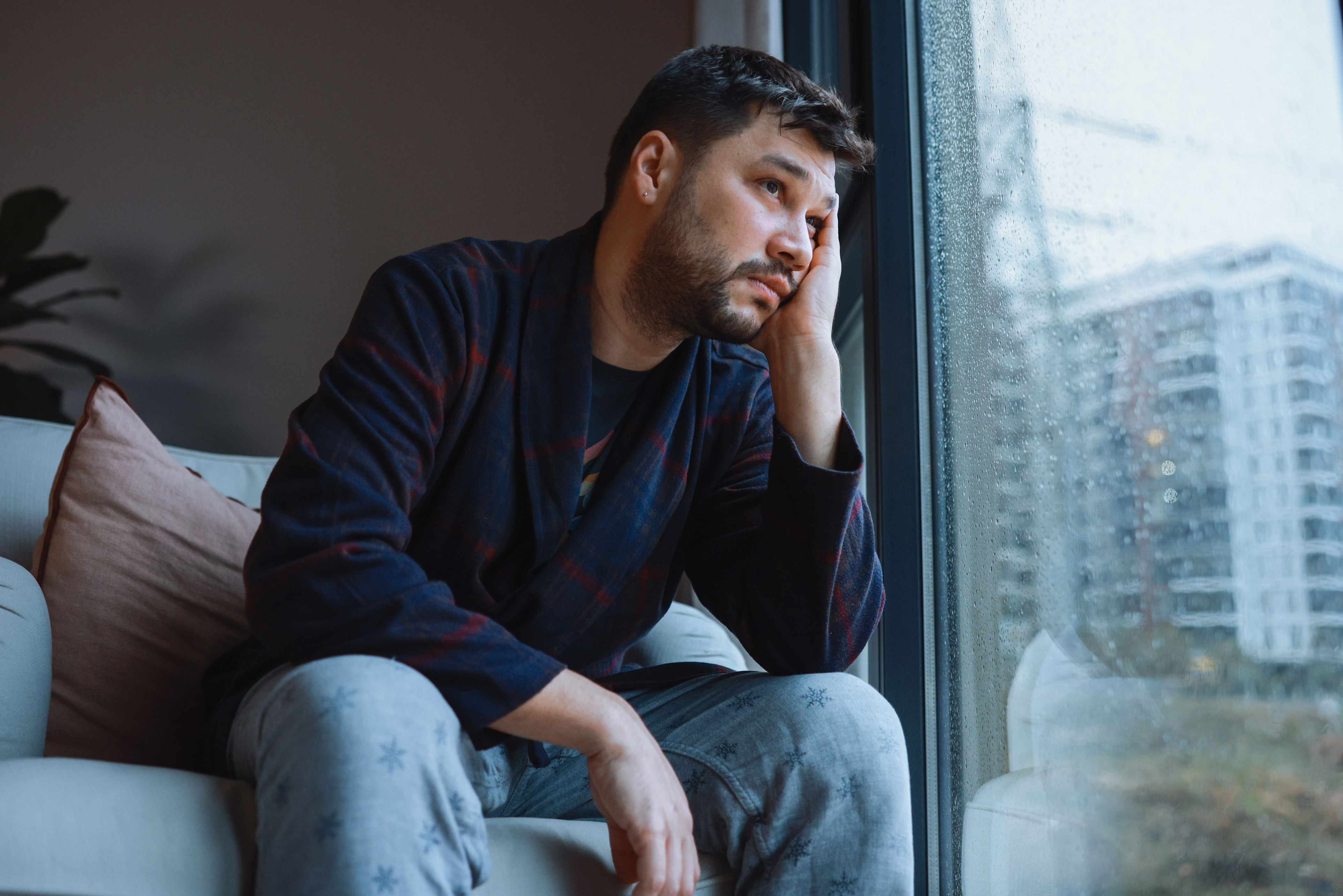 A person looks thoughtful while sitting on a chair near a window with raindrops outside, wearing casual loungewear at home