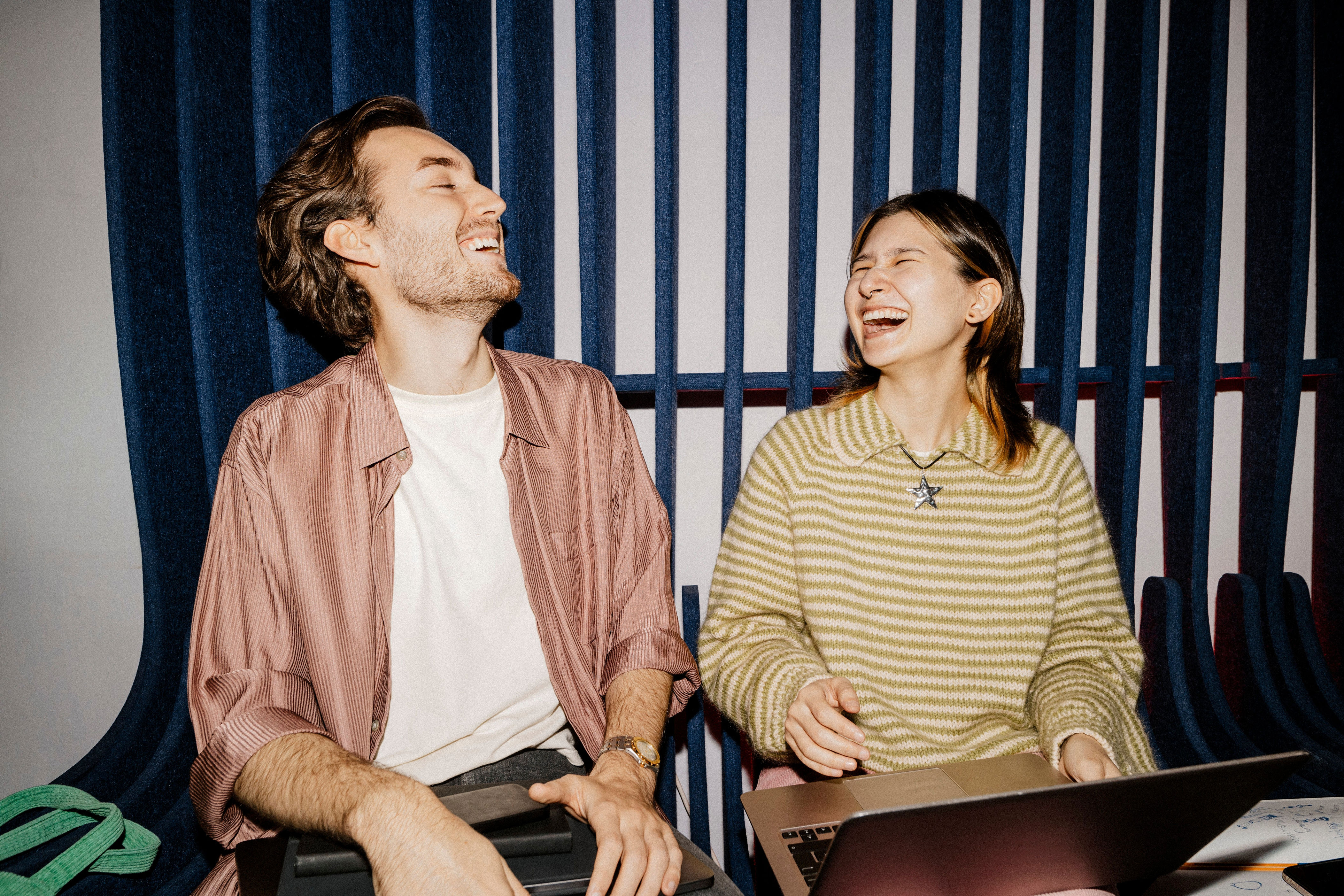 Two people laugh together while sitting on a bench, one holding a laptop. They seem to be enjoying a lighthearted moment