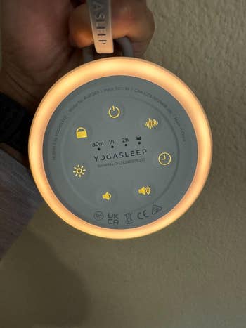 Person holding a Yogasleep sound machine featuring timer and volume icons, symbolizing easy control for customizable sleep aid settings