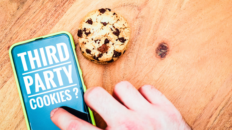 A mobile phone next to a cookie on a wooden table with its screen reading Third Party Cookies