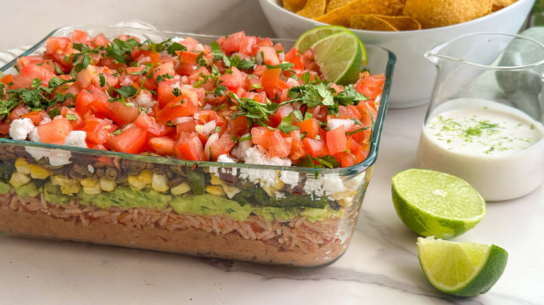7-layer dip in rectangular glass dish, next to jug of crema and fresh lime