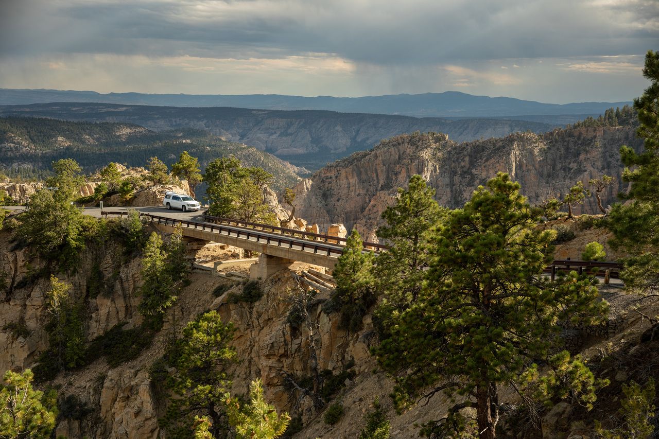 A vehicle crossing a bridge over a deep canyon with surrounding forest and distant mountains