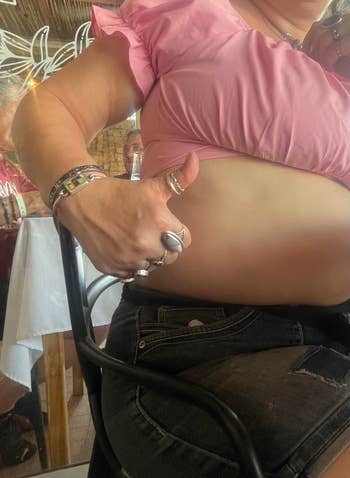 Person in casual attire showing thumb under shirt, revealing abdomen. Various rings and bracelets worn. Indoor setting with tables in the background