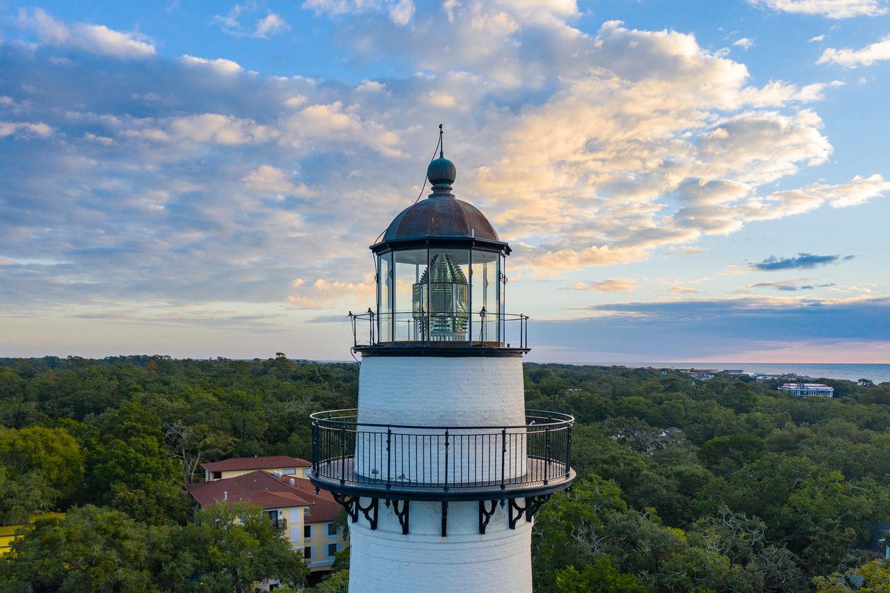 A lighthouse rises above a wooded area under a cloudfilled sky with glimpses of buildings and the horizon in the distance