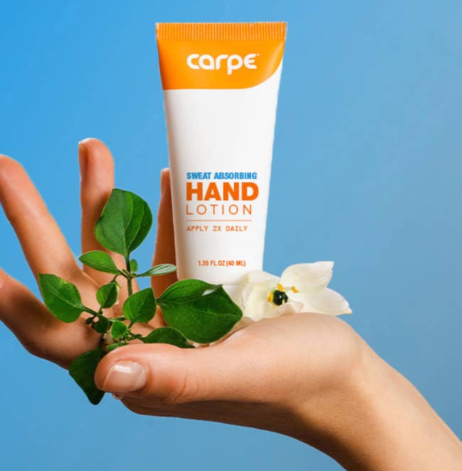 A hand holds a tube of Carpe sweat-absorbing hand lotion with a small plant and flower, suggesting freshness and natural ingredients