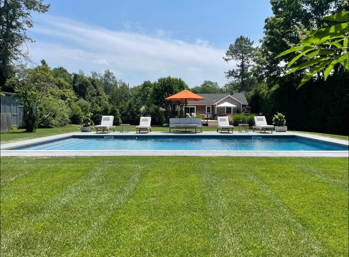 Backyard swimming pool with lounge chairs, orange umbrella, lawn, trees, and a small house in the background.