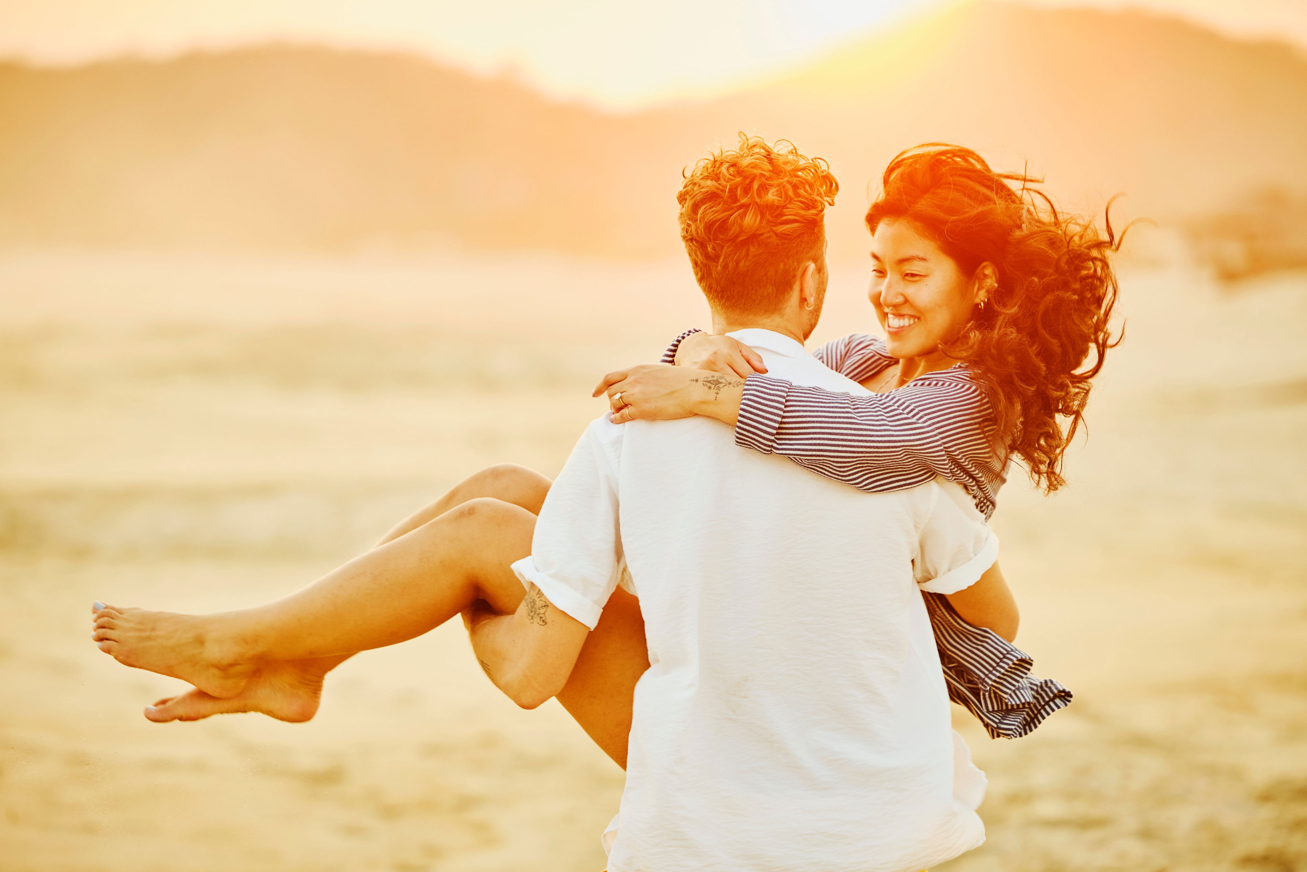 A person happily holds another in their arms on a sunlit beach, both smiling and enjoying the moment