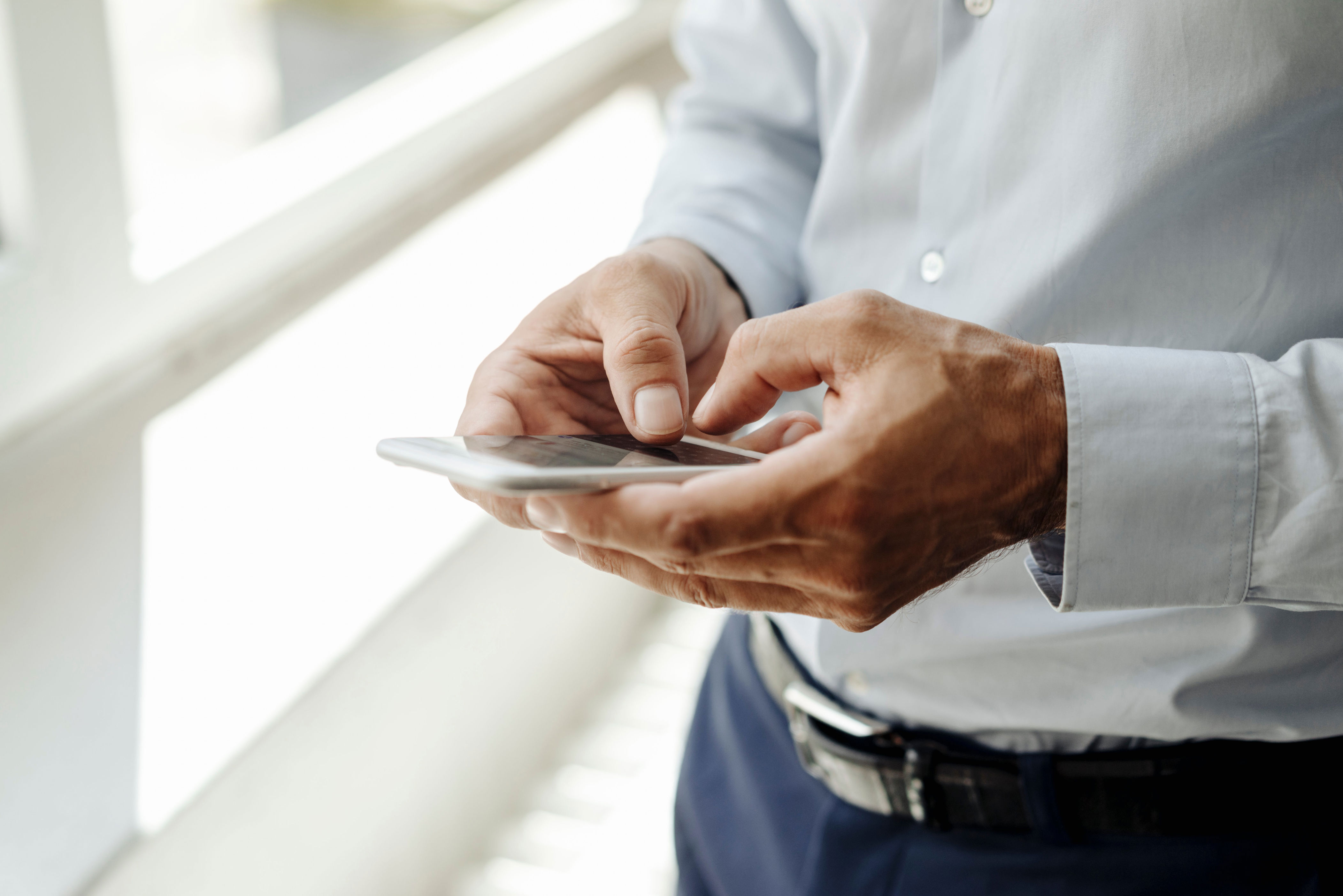 Person in a business shirt using a smartphone by a window, focusing on the screen with both hands