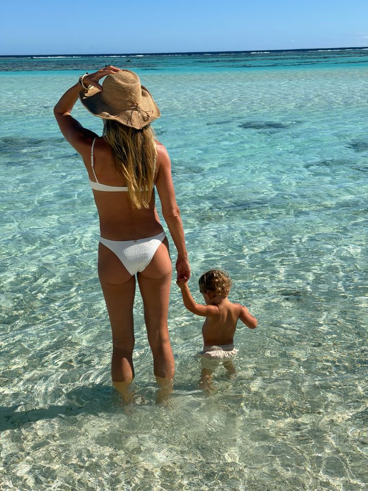 Gisele Bündchen holds her one-year-old son's hand as they stand in clear blue water along the shore. Bündchen is wearing a white bikini and a tan sunhat, while her son is wearing khaki swim trunks.