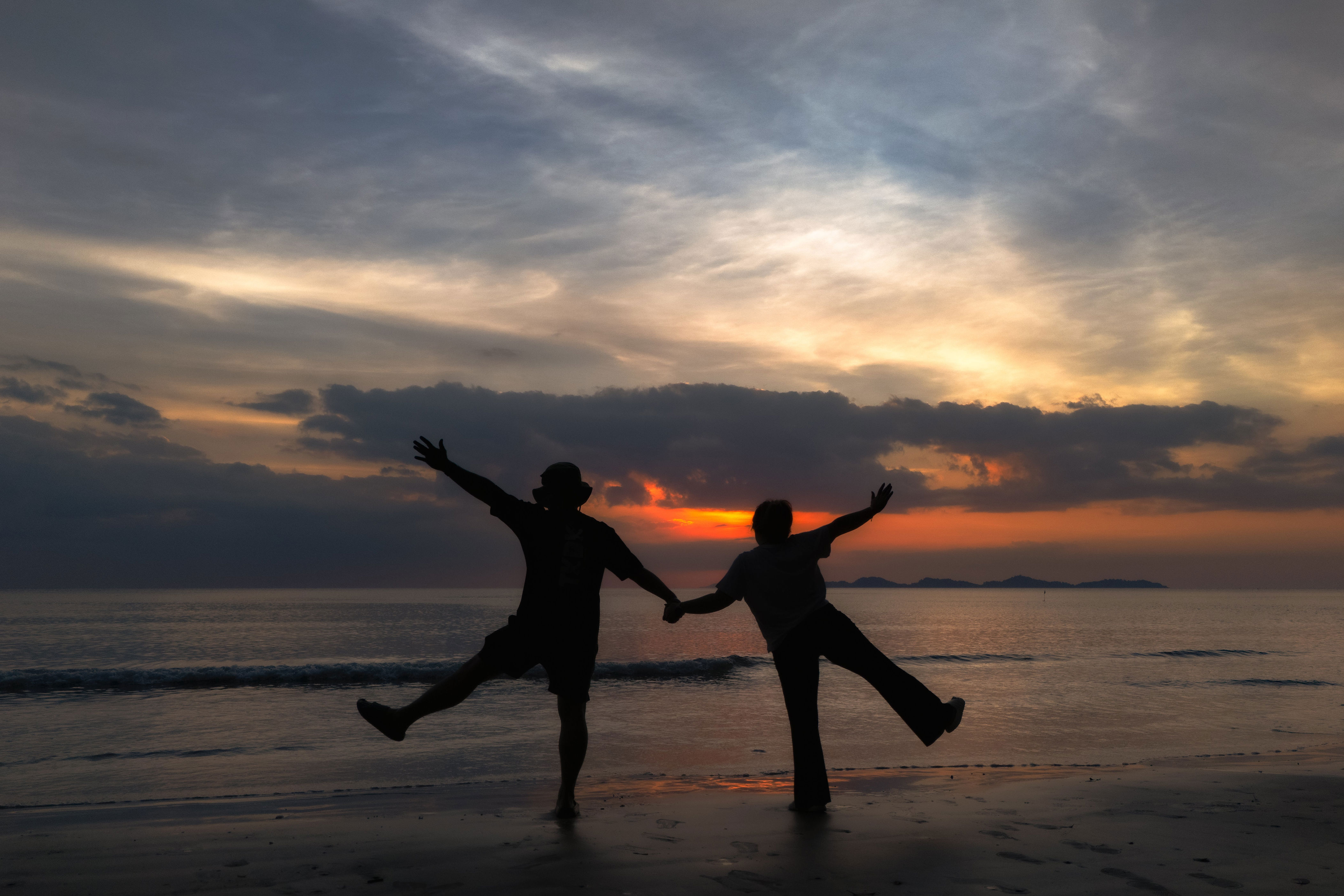 Two people holding hands, playfully silhouetted against the sunset on a beach, evoking a sense of romance and joy