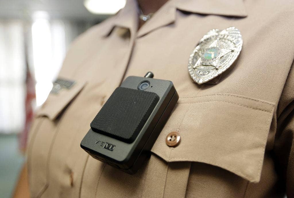 A close-up of a police officer's uniform shows a body-worn camera clipped to the front of the shirt beneath a law enforcement badge.