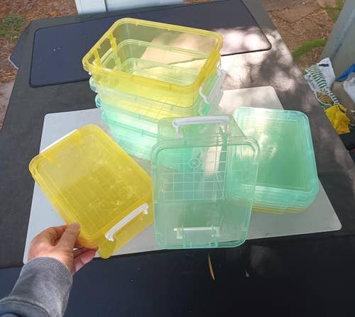 Person holding a stack of transparent plastic storage containers with white handles outdoors on a table