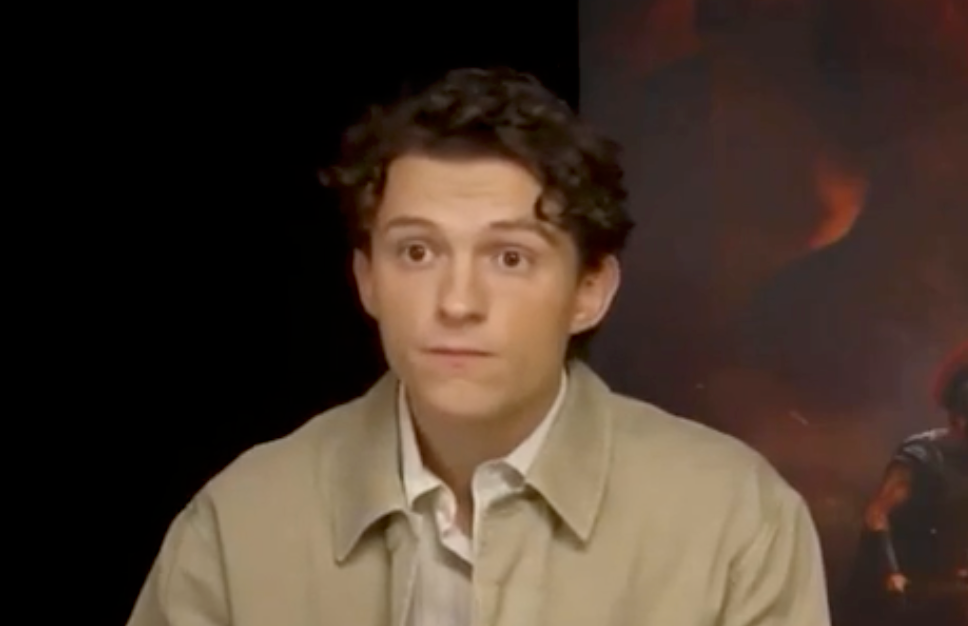Person in a beige jacket and white shirt, sitting against a dark background, looking forward with a neutral expression