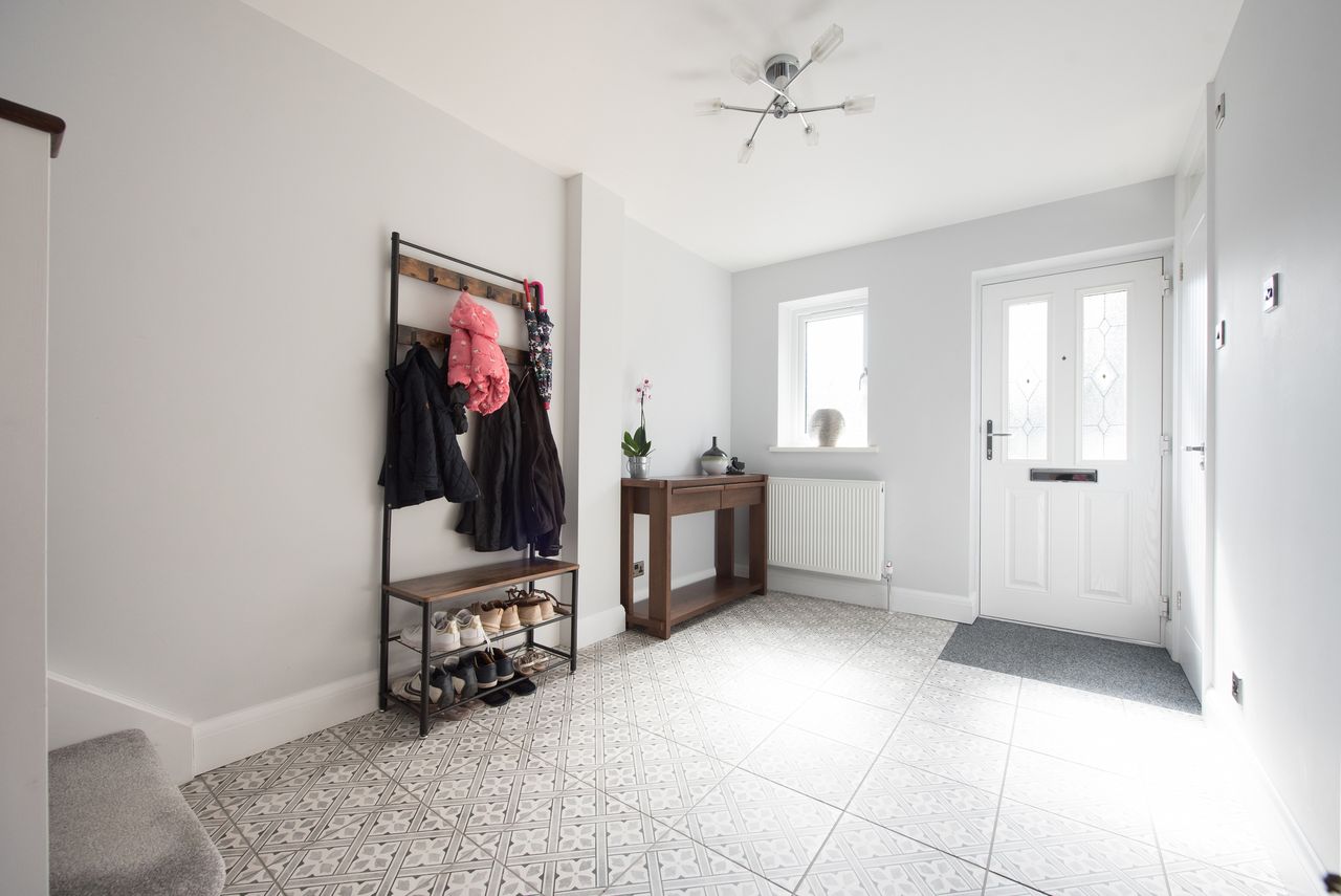 A general view of a hall with patterned tiled floor with coats hanging on hooks and shoes on shelves and console table within a home