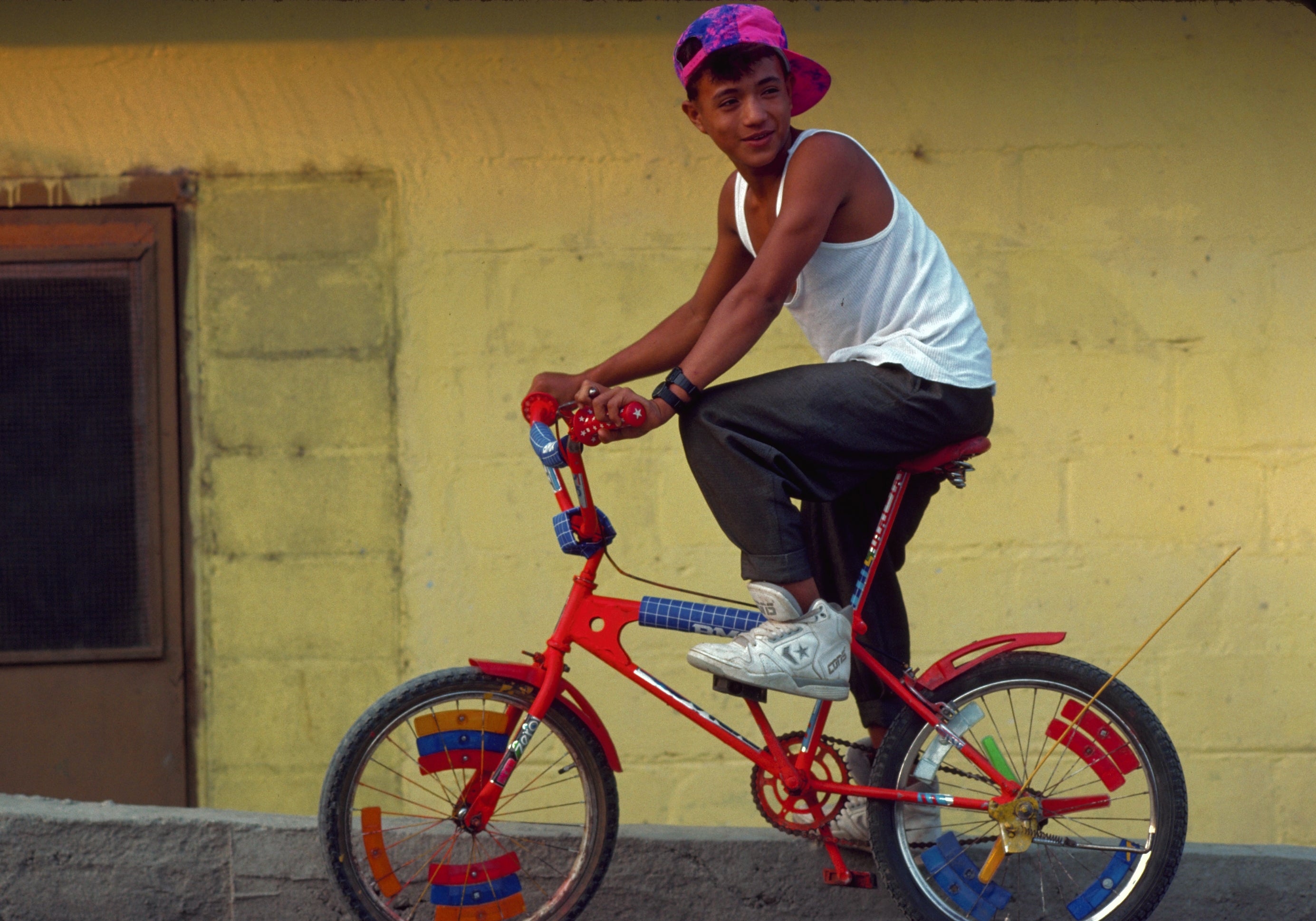 A young person in casual attire, white tank top and colorful cap, rides a bright, customized bicycle on a sloped street next to a building