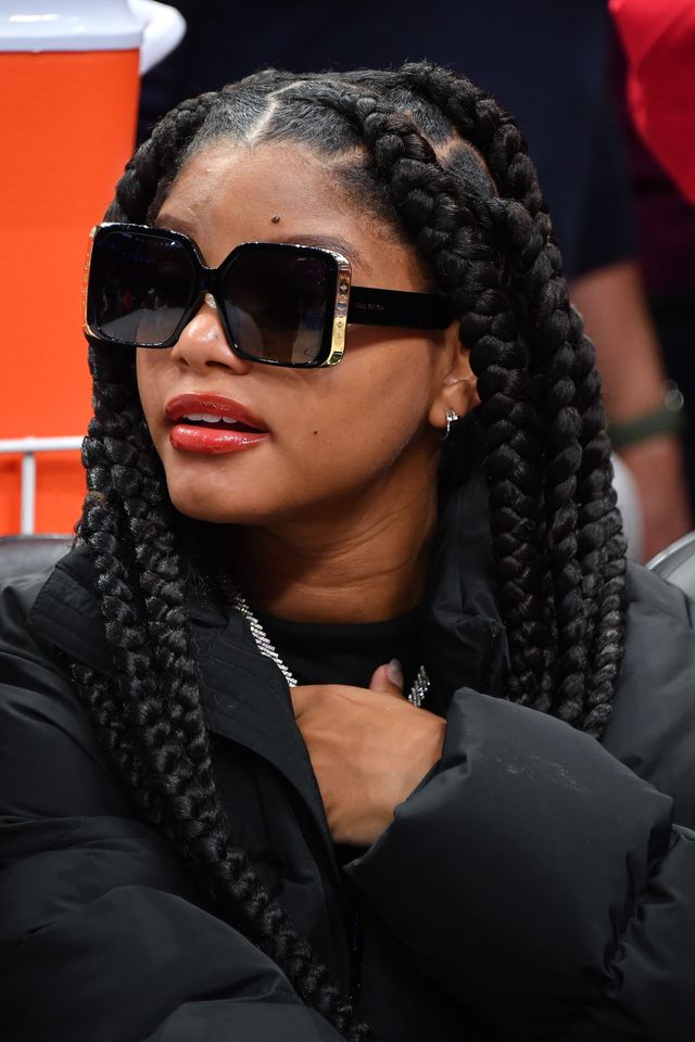 A person with braided hair wearing sunglasses sits in an indoor setting