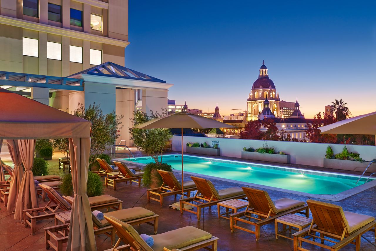 A rooftop swimming pool with lounge chairs and umbrellas with a cityscape including a prominent dome in the background during sunset