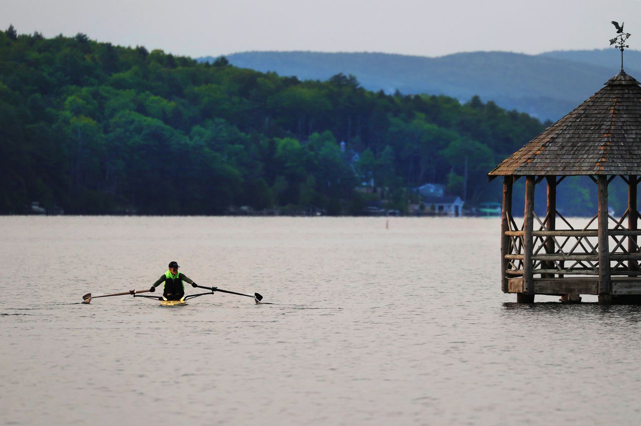 Person rowing a kayak on a lake near a wooden gazebo with a forested shoreline in the background captured at Lake Winnipesaukee New Hampshire