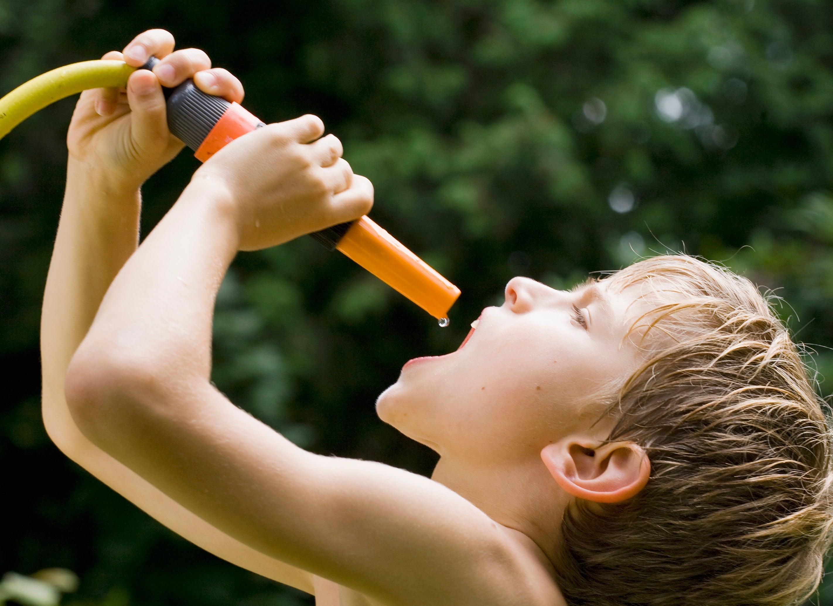 Child drinks water from a garden hose outdoors, surrounded by greenery
