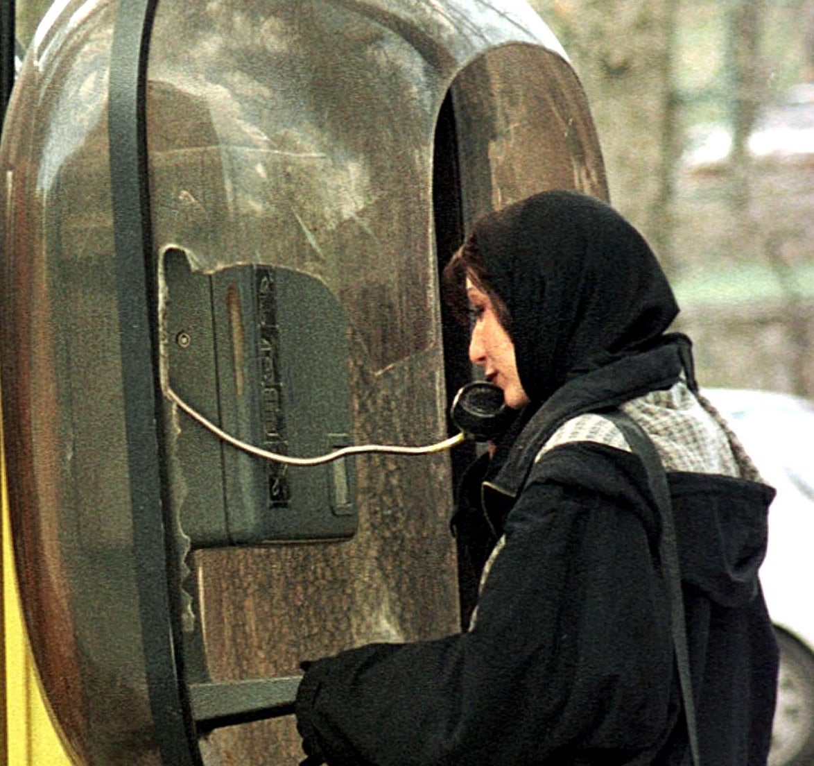 Person using a public phone booth, wearing a hoodie, jeans, and platform shoes, standing on a street