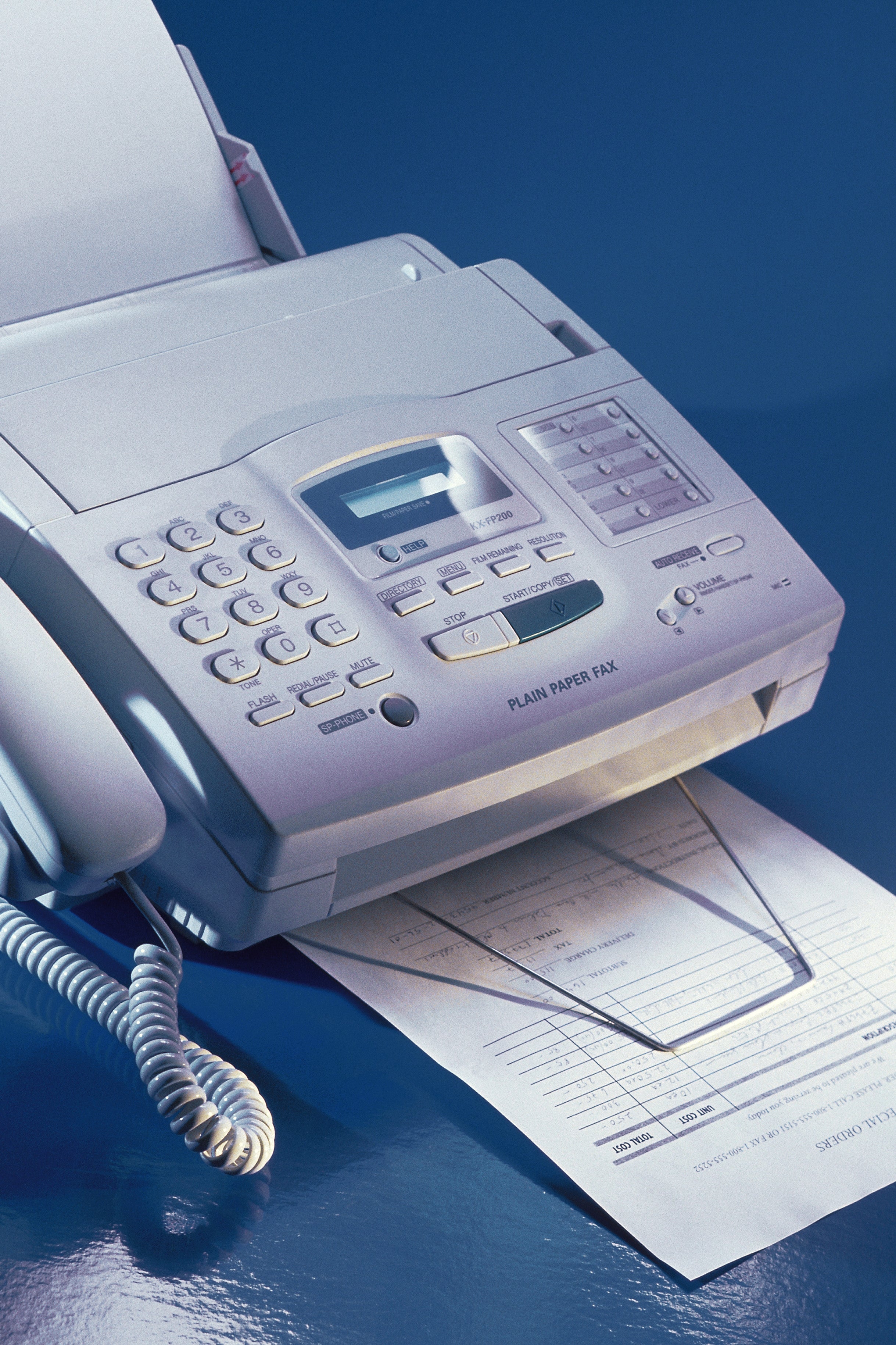 Fax machine on a desk, paper emerging from the tray, with coiled phone handset attached