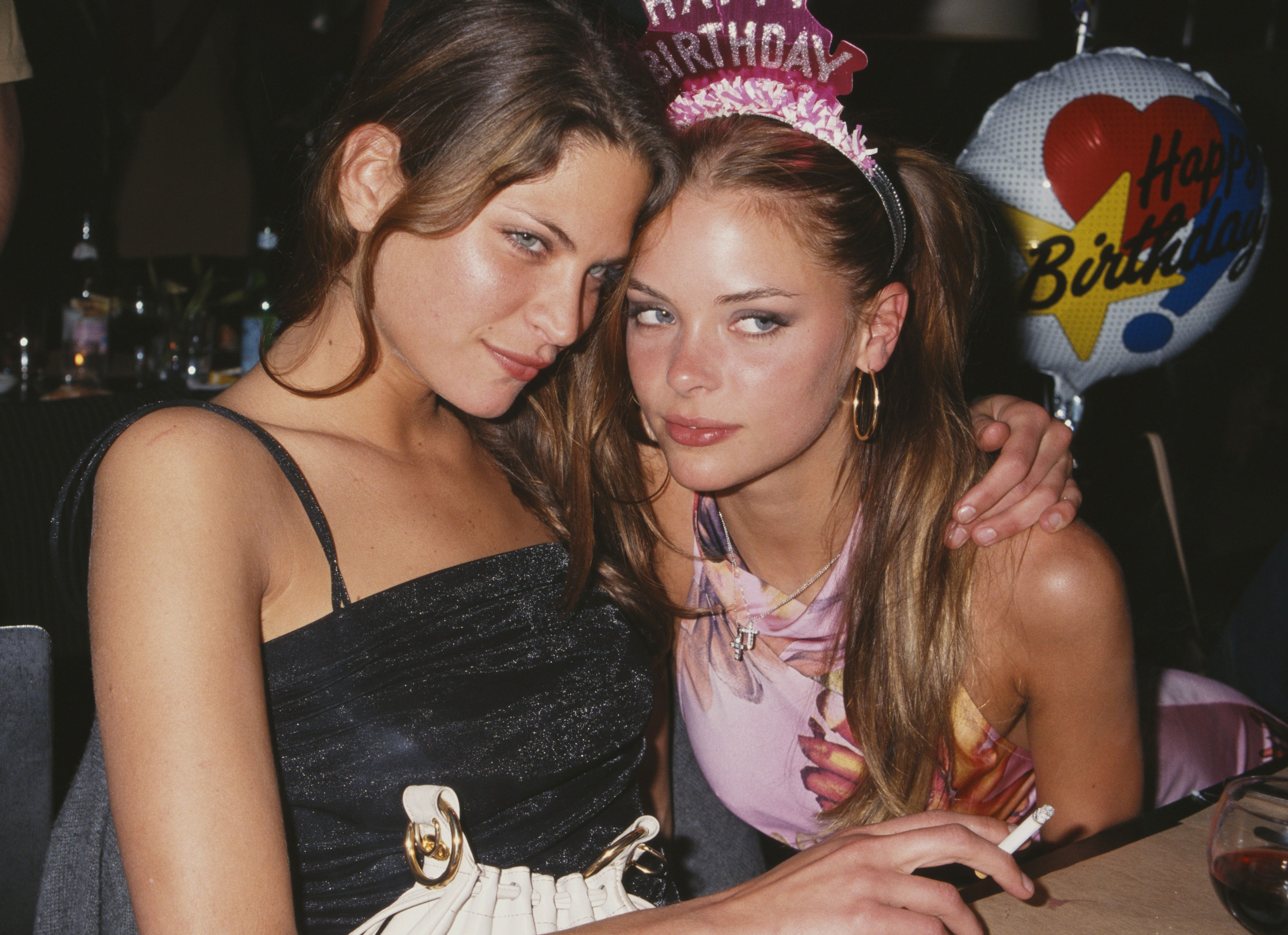 Two friends celebrating a birthday, one wearing a "Happy Birthday" headband, at a party. They pose closely with party decor in the background