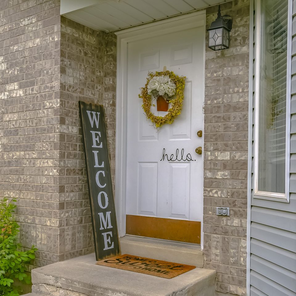 A front door with a decorative wreath a mat and a vertical Welcome sign placed nearby