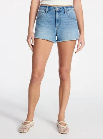 Person wearing a pair of mid-thigh, frayed hem denim shorts and mesh ballet flats