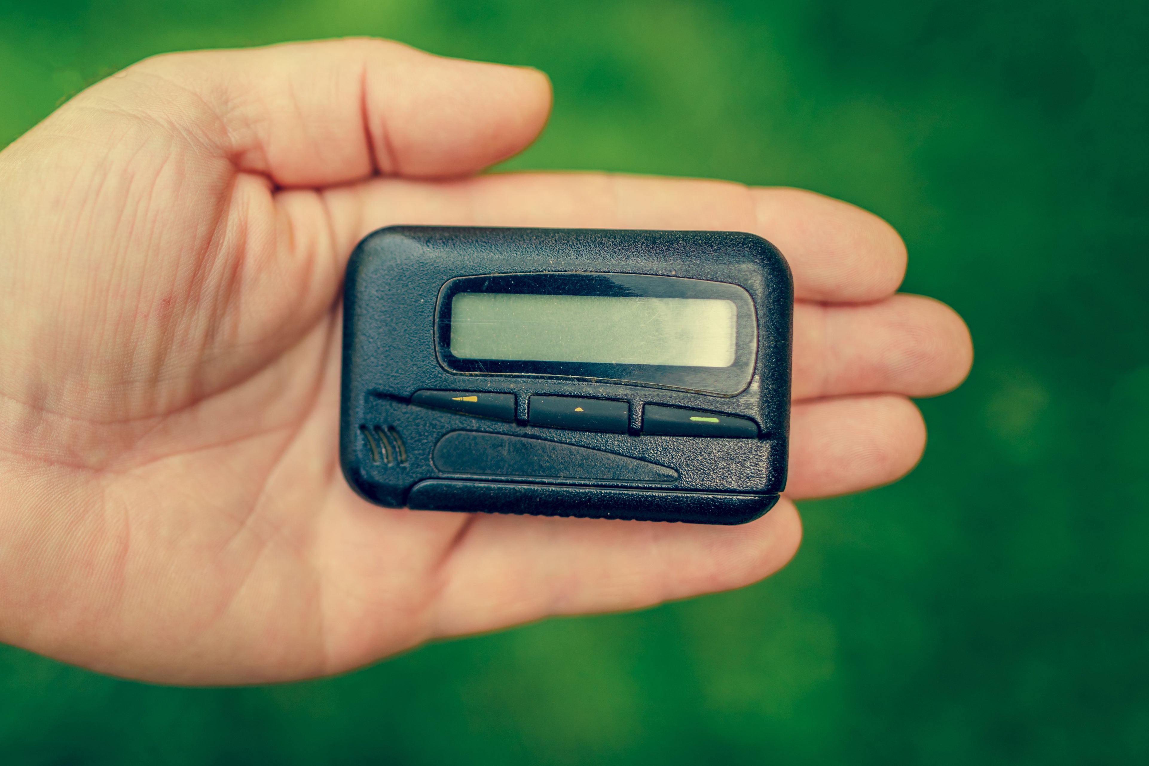 Hand holding a small, rectangular electronic pager with a simple display and buttons, against a blurred green background