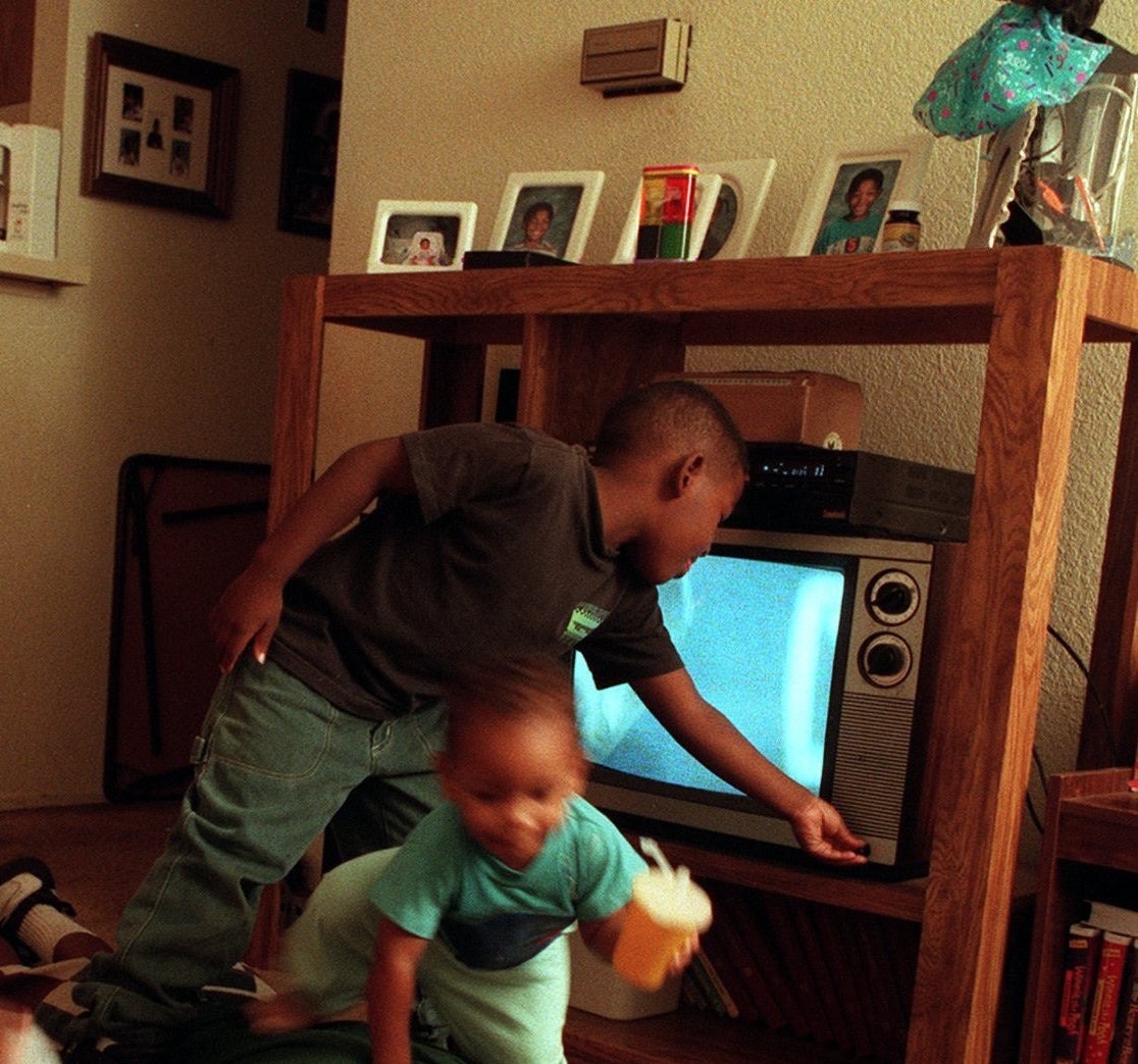 Two young children play in a living room. One child looks at a TV set while the other holds a cup. Family photos are on a shelf behind them