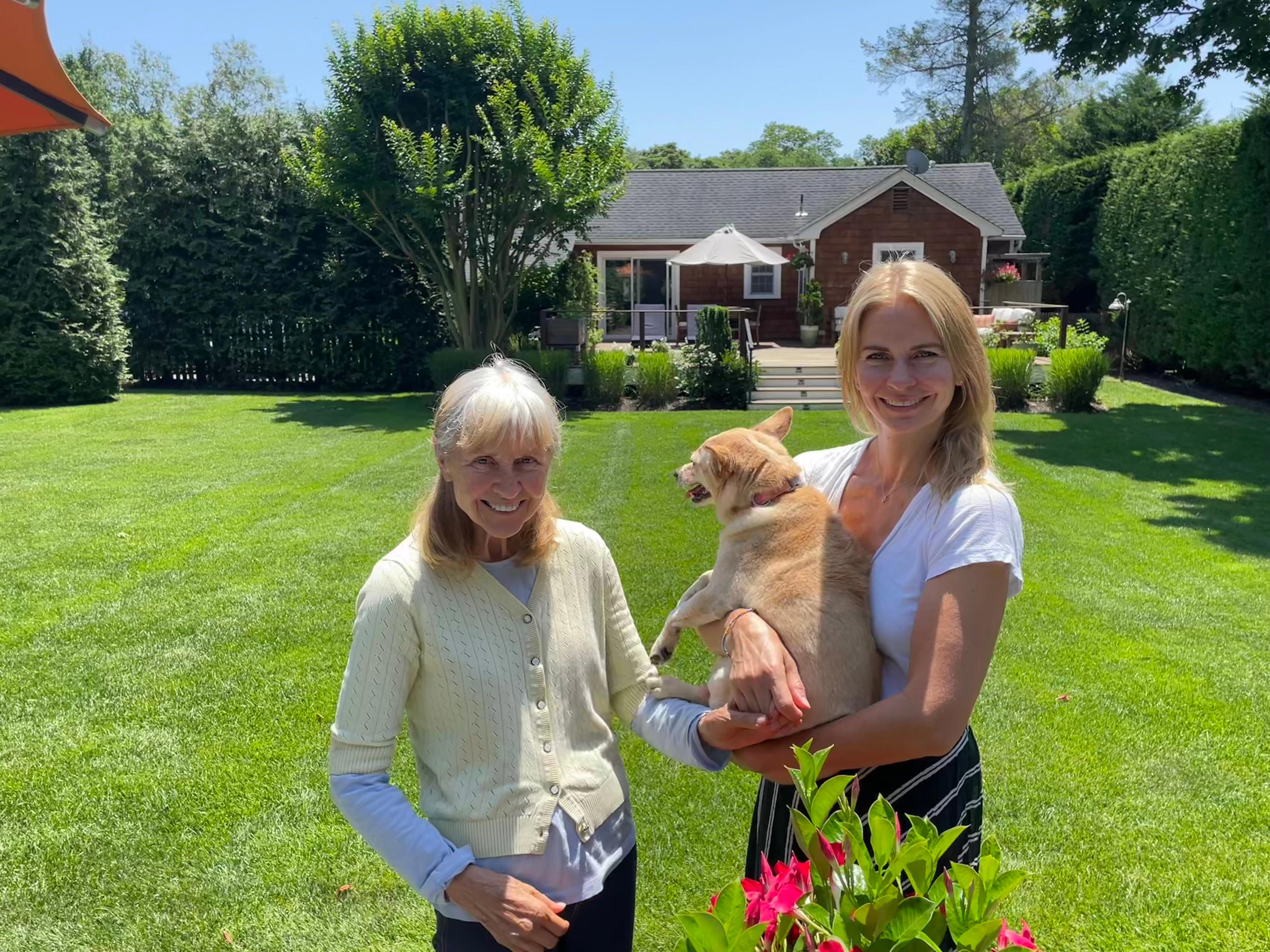 Two people stand on a sunny backyard lawn, one holding a small dog, with a house and garden behind them.