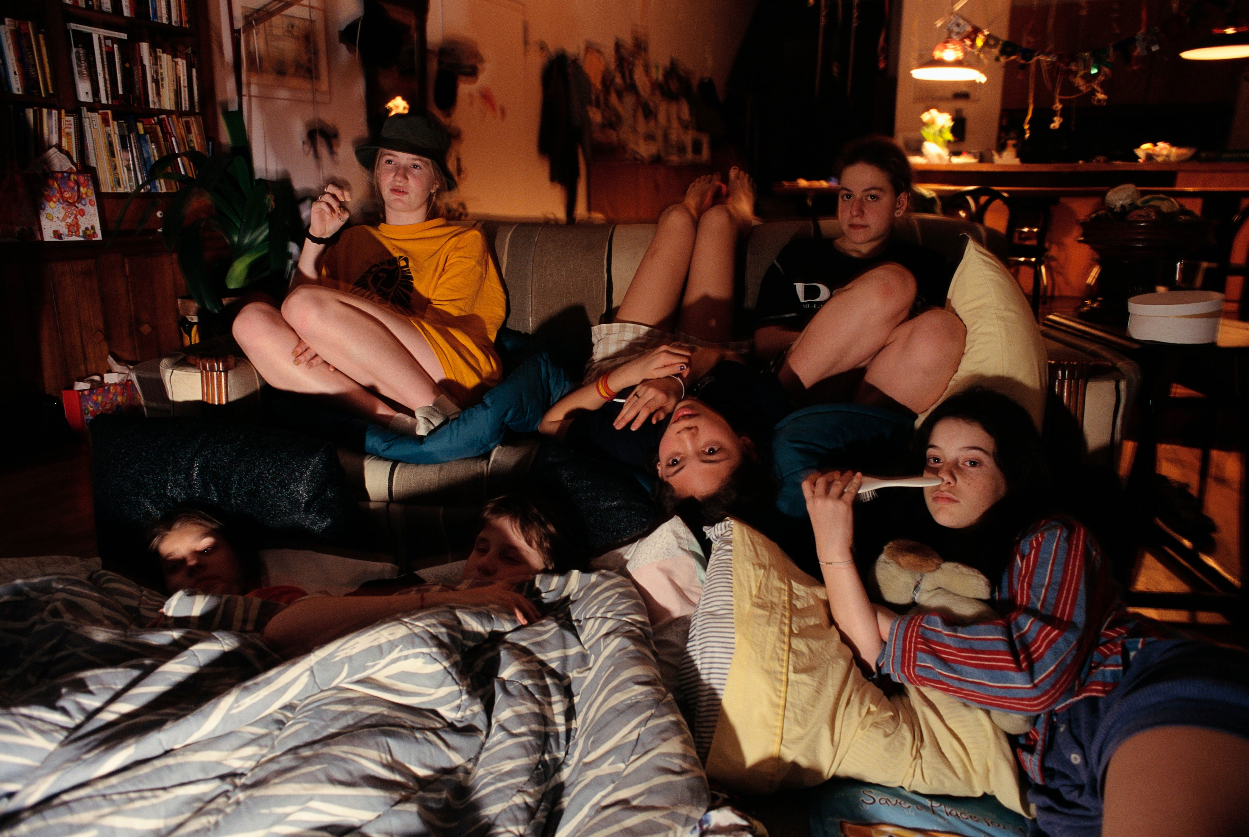 Group of people lounging on a sofa and floor in a cozy room, watching TV, surrounded by pillows and blankets
