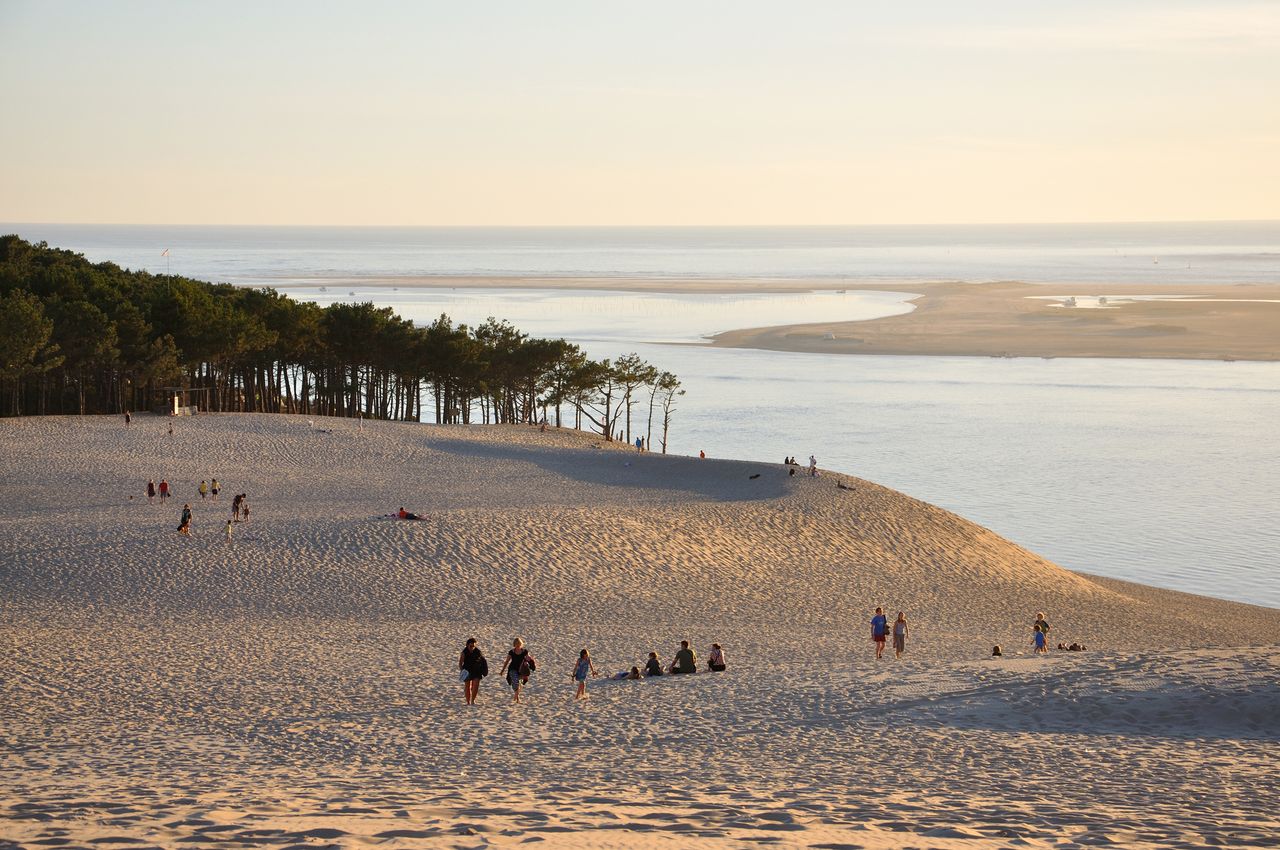 A scenic view of sand dunes with sparse vegetation and people walking overlooking water and distant land masses